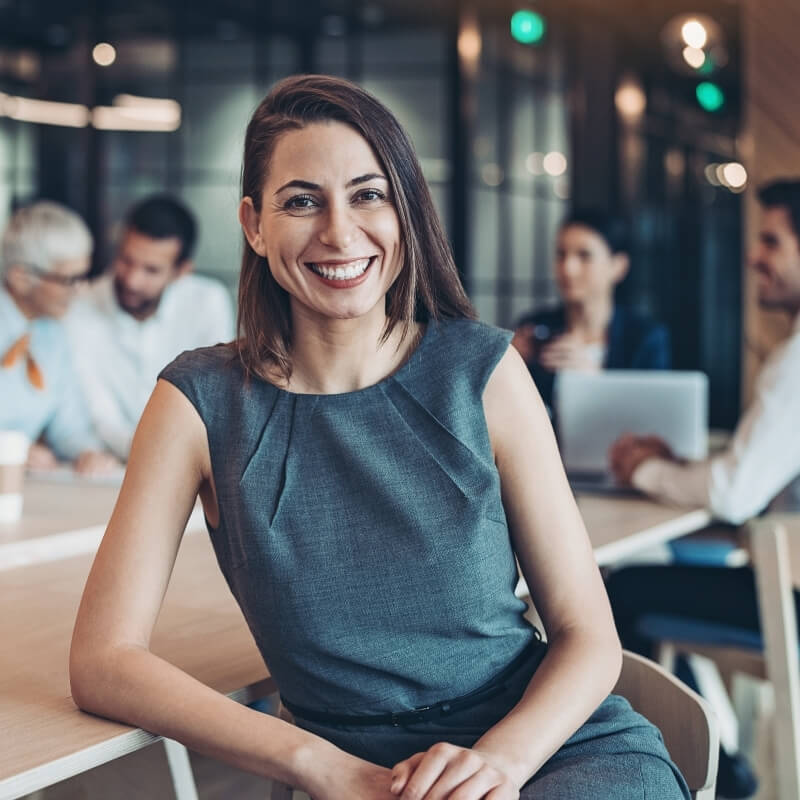 female leader smiling leaning on desk, people working behind her